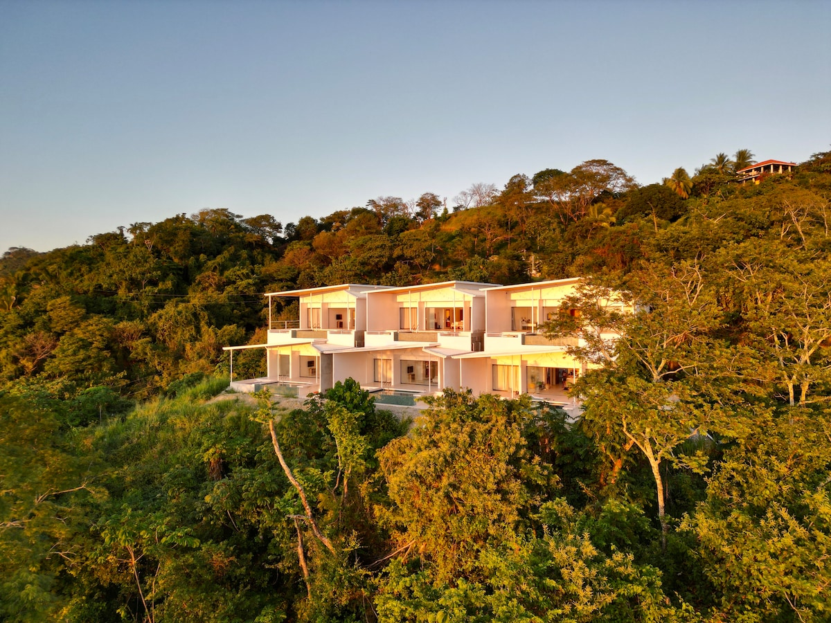 The exterior of Casa Topaze is surrounded by lush greenery, showcasing a modern design with multiple levels. Large glass doors and windows are visible, allowing natural light and offering expansive views of the surrounding jungle. The building is integrated into the hillside, enhancing its serene setting.