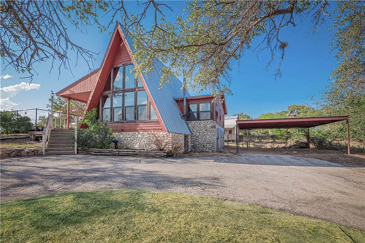 The A-frame cabin features a striking triangular shape, highlighted by large windows that create an open and airy feel. A stone foundation adds rustic charm, while a covered parking area is visible on the side. Lush green grass surrounds the property, adding to the natural setting.