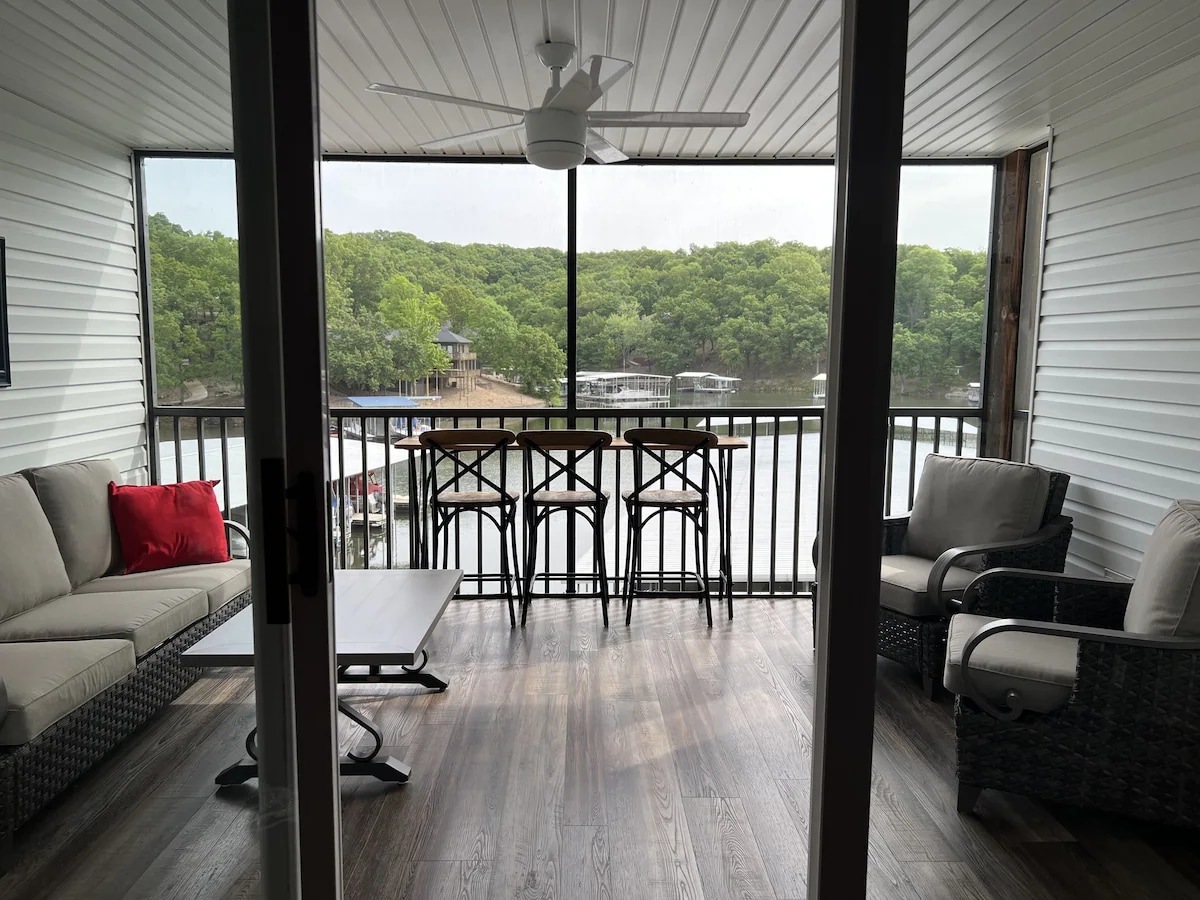 A covered balcony offers a view of the water surrounded by greenery. Seating includes a sectional sofa and a tall table with chairs, providing a space for relaxation or dining. Natural light filters through large sliding doors, enhancing the open and airy atmosphere.