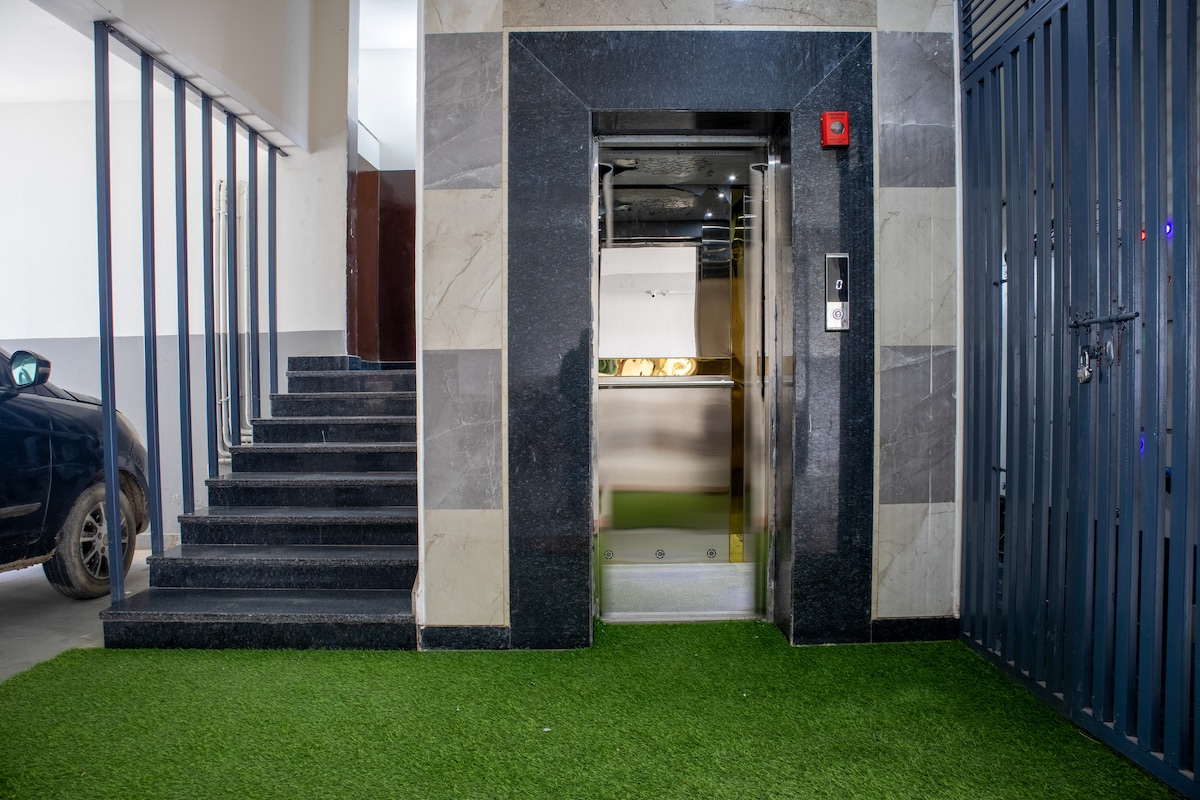 An elevator is positioned beside a staircase, both framed by sleek, polished marble finishes. The entrance is enhanced by artificial grass, providing a refreshing touch. The environment is modern, combining functionality with design elements that create an accessible entry point.