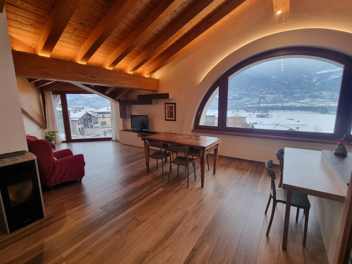 A spacious living area features large arched windows offering expansive views of the surrounding mountains and snow-covered scenery. Wooden beams create a warm atmosphere, complemented by natural light. A dining table and chairs are present, along with a cozy red sofa and a television for relaxation.
