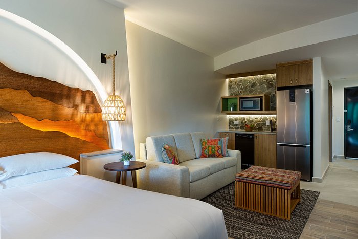A bright studio layout is highlighted by a spacious bed and a comfortable sofa bed. A modern kitchenette is visible, along with a textured accent wall that adds warmth. Natural light filters through the room, creating a welcoming ambiance for guests.