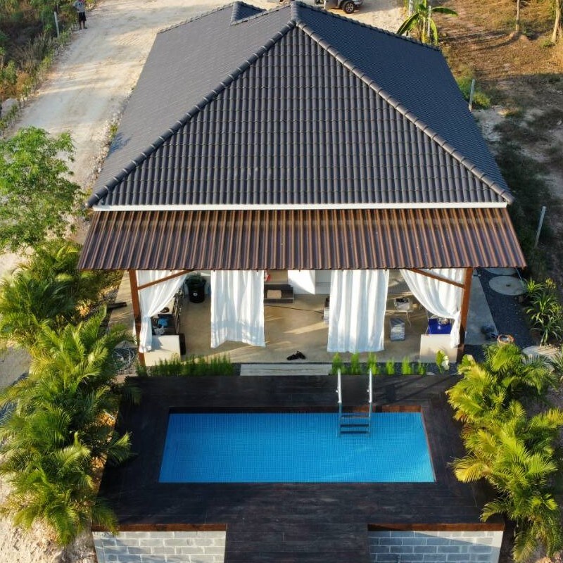 An aerial view of a small house features a sloped roof with dark tiles. An inviting swimming pool is positioned on a wooden deck, surrounded by greenery. Flowing white curtains frame the entrance, enhancing the open and airy feel of the space.