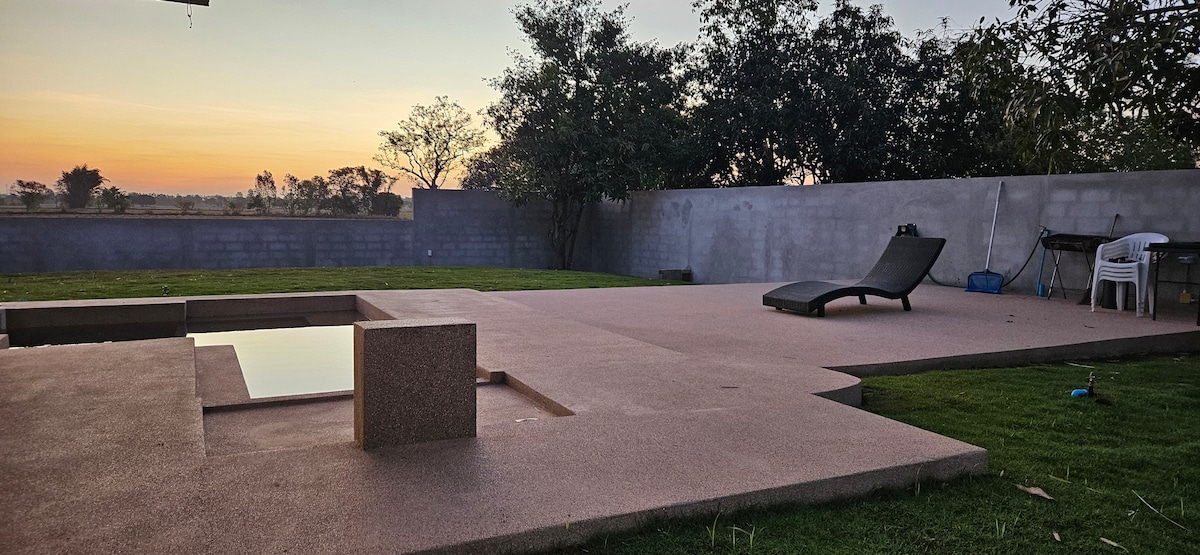 An outdoor area features a sleek pool surrounded by a textured stone surface. A lounge chair is positioned nearby, with the backdrop of lush greenery and a softly lit sky at sunset.