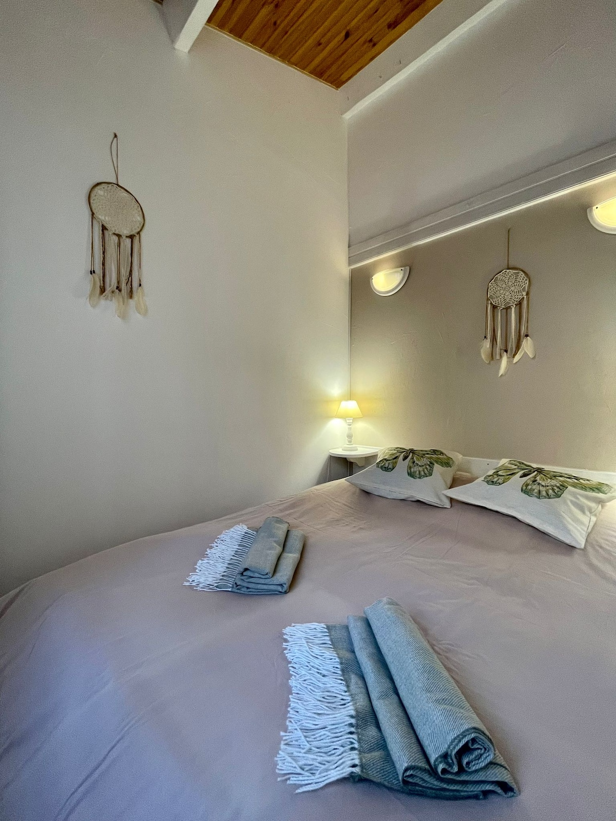 A cozy sleeping area features a spacious bed dressed in soft linens. Dream catchers hang on the walls, complemented by ambient lighting. Towels are neatly arranged at the foot of the bed, creating a welcoming and organized appearance.