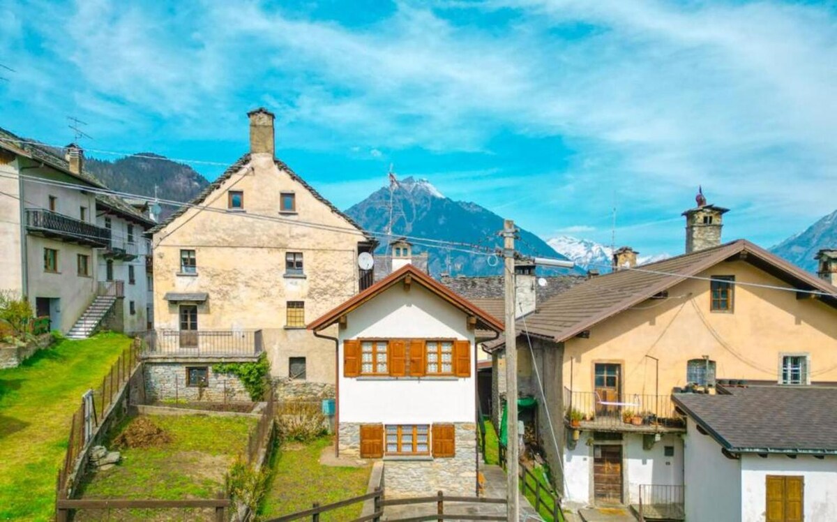 A charming village setting features a collection of traditional alpine buildings, showcasing a mix of stone and wood façades. Mountain peaks rise in the background under a clear blue sky, while lush green lawns and well-maintained gardens add a refreshing touch to the landscape.
