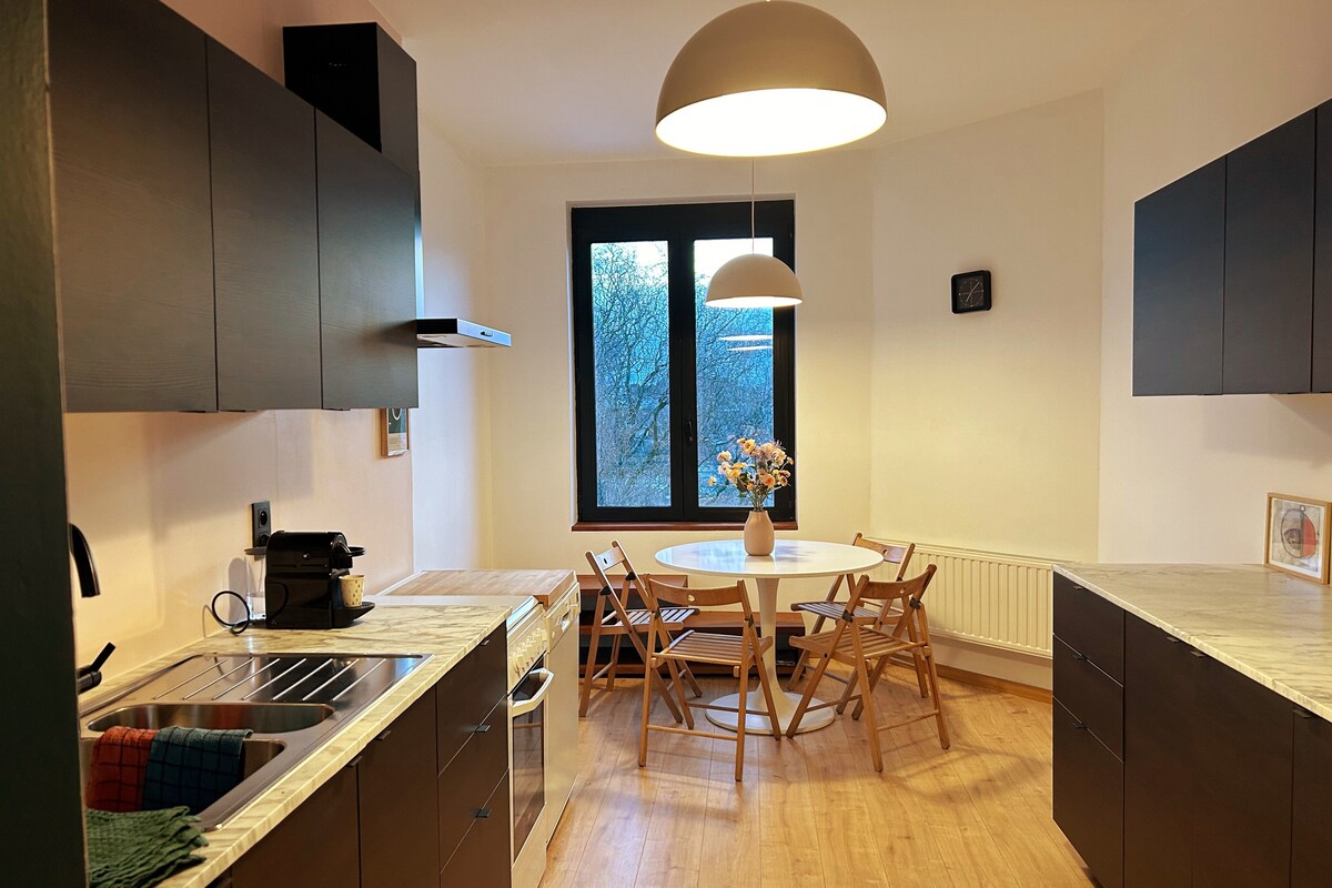 The kitchen space features dark cabinetry with a marble countertop and stainless steel appliances. A round dining table with folding chairs is positioned near a large window, allowing natural light to brighten the room. A small vase with flowers adds a touch of color.