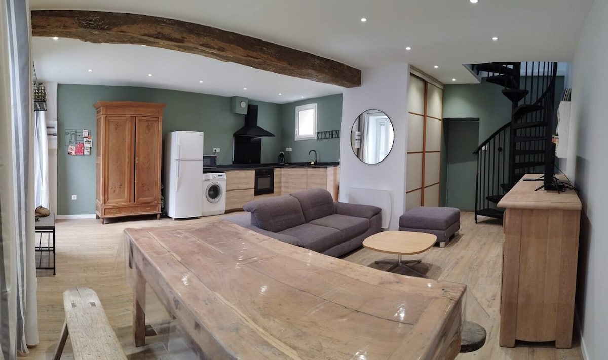 An open living area is presented, featuring a mix of natural wood elements and modern design. A spacious sofa faces a minimalist entertainment unit, and a large wooden dining table is positioned centrally. A spiral staircase leads to the upper level, while large windows allow natural light to fill the room.