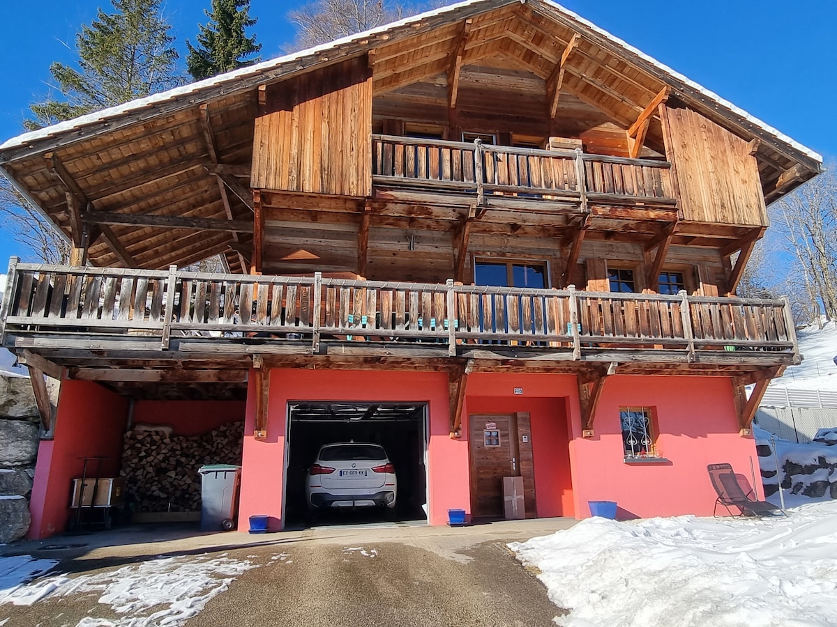 A charming wooden chalet is depicted, featuring a spacious balcony with wooden railings. The lower level has a garage with a white vehicle parked in front. The exterior is painted in a warm pink hue, contributing to the inviting presence against a snowy backdrop.