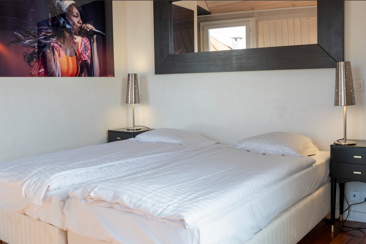 A spacious guest room features a large bed with crisp white bedding. Two bedside tables equipped with lamps are positioned on either side. A large mirror hangs above the bed, reflecting light from the nearby window, which is framed by a minimalist design.