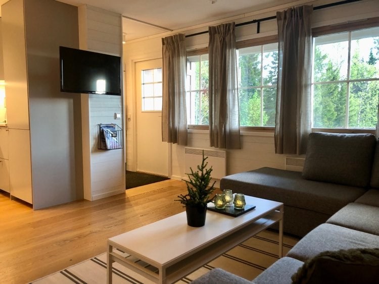 A bright living area features large windows adorned with light curtains, allowing natural light to fill the space. A comfortable sectional sofa is positioned around a low coffee table, complemented by decorative candles. A television is mounted on the wall opposite the entrance.