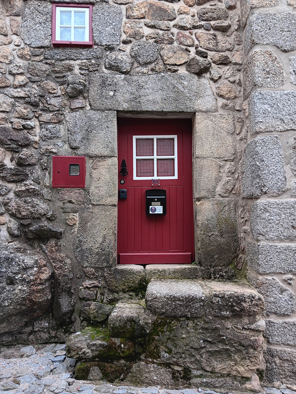 A rustic entrance features a red wooden door with a grid-patterned window, set against a stone wall. A black mailbox is mounted next to the door, and a small light fixture adds subtle illumination. Stone steps lead up to the entrance, lined with moss.
