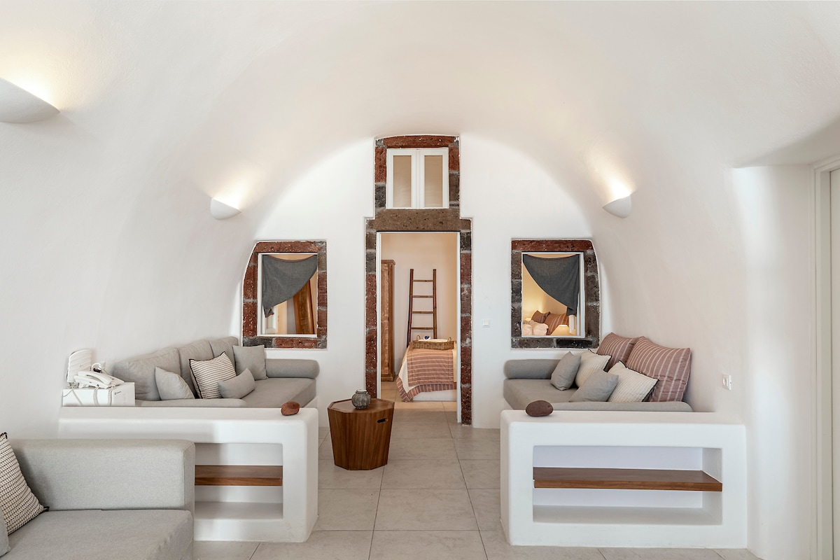 A spacious living area showcases soft, light-colored seating arrangements. Natural light streams through the arched windows, illuminating the neutral-toned decor. Textured walls and simple wooden accents enhance the serene atmosphere, while a small table sits centrally, creating an inviting space for relaxation.