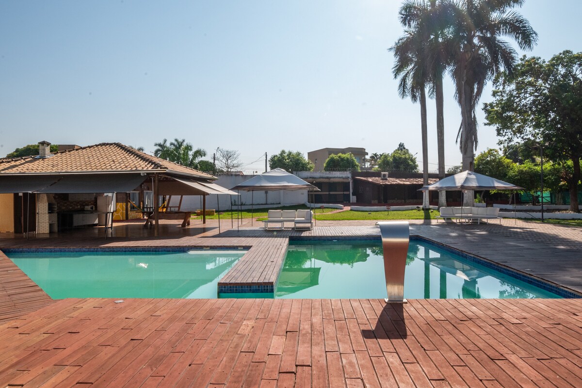 The pool area features a large swimming pool framed by wooden decking, surrounded by greenery and several palm trees. A shaded outdoor area with tables and umbrellas is visible, along with a barbecue kiosk in the background. Sunlight reflects off the water, creating a serene atmosphere.