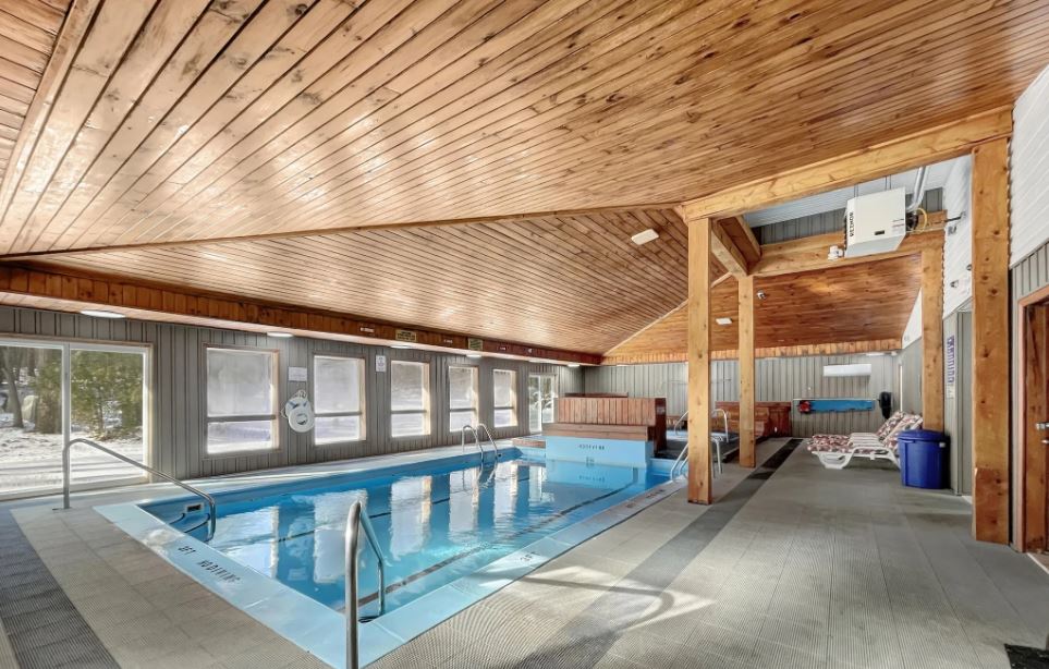 An indoor pool area is featured, with a wooden ceiling and large windows allowing natural light. The clear blue pool is flanked by safety rails, and lounge chairs are arranged along the sides. The space is inviting and well-maintained.