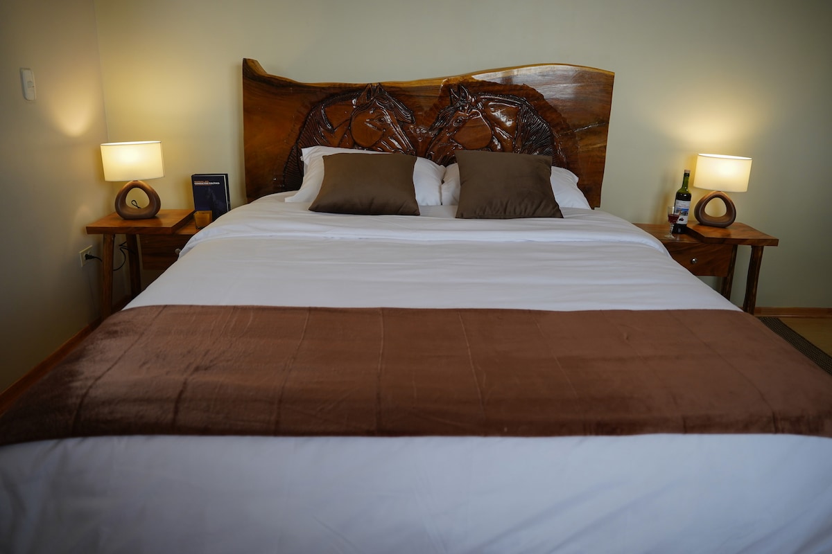 A spacious bed is framed by intricately carved wooden headboard featuring horse motifs. Two bedside tables hold soft lamps, and decorative pillows in earthy tones add comfort. A bottle of wine rests on one table, suggesting a cozy atmosphere for relaxation.