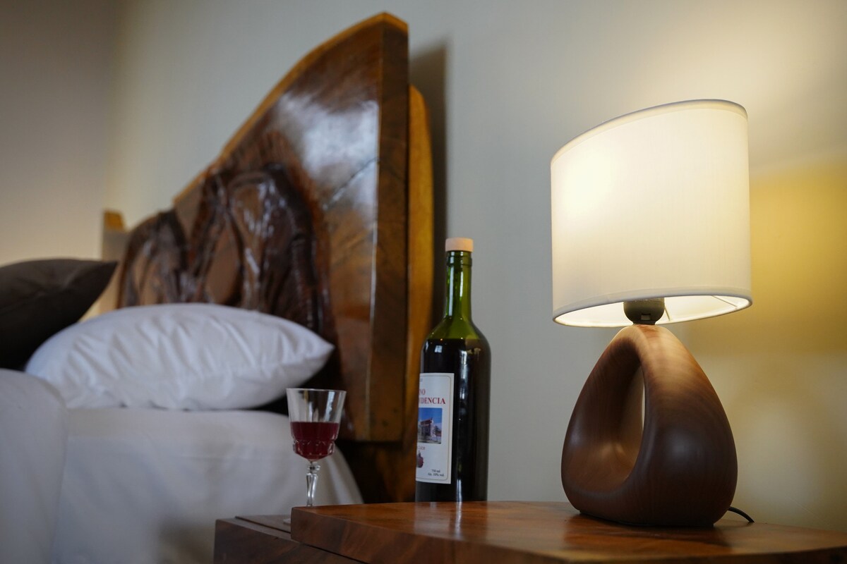 A bedside table is displayed with a unique lamp and a bottle of wine, accompanied by a glass of red liquid. The headboard, crafted from natural wood, features intricate carvings, providing an elegant backdrop to the cozy arrangement.