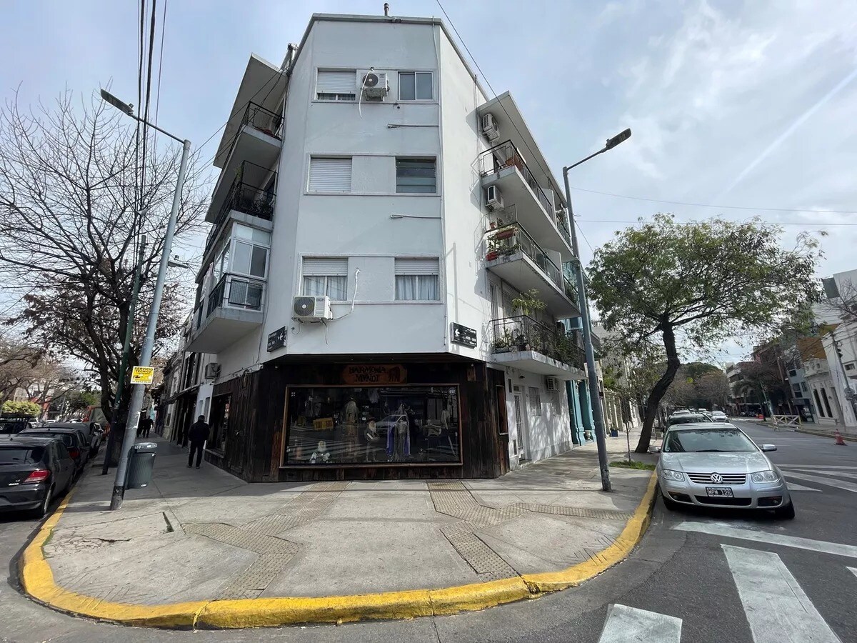 The exterior of a modern multi-story building is shown, featuring balconies adorned with plants. Large windows provide ample light, and a retail space occupies the ground floor. Nearby, trees and street signage complete the urban scene, highlighting the neighborhood's vibrant atmosphere.