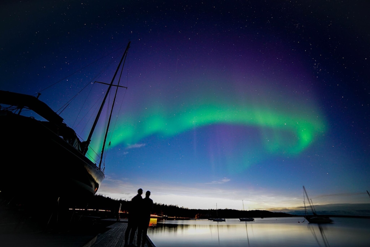 The vibrant Northern Lights arc gracefully over a calm waterway, reflecting hues of green and purple. Silhouettes of two individuals stand on a dock, framed by boats moored nearby. The twilight sky transitions from deep navy to soft azure, creating a serene backdrop.