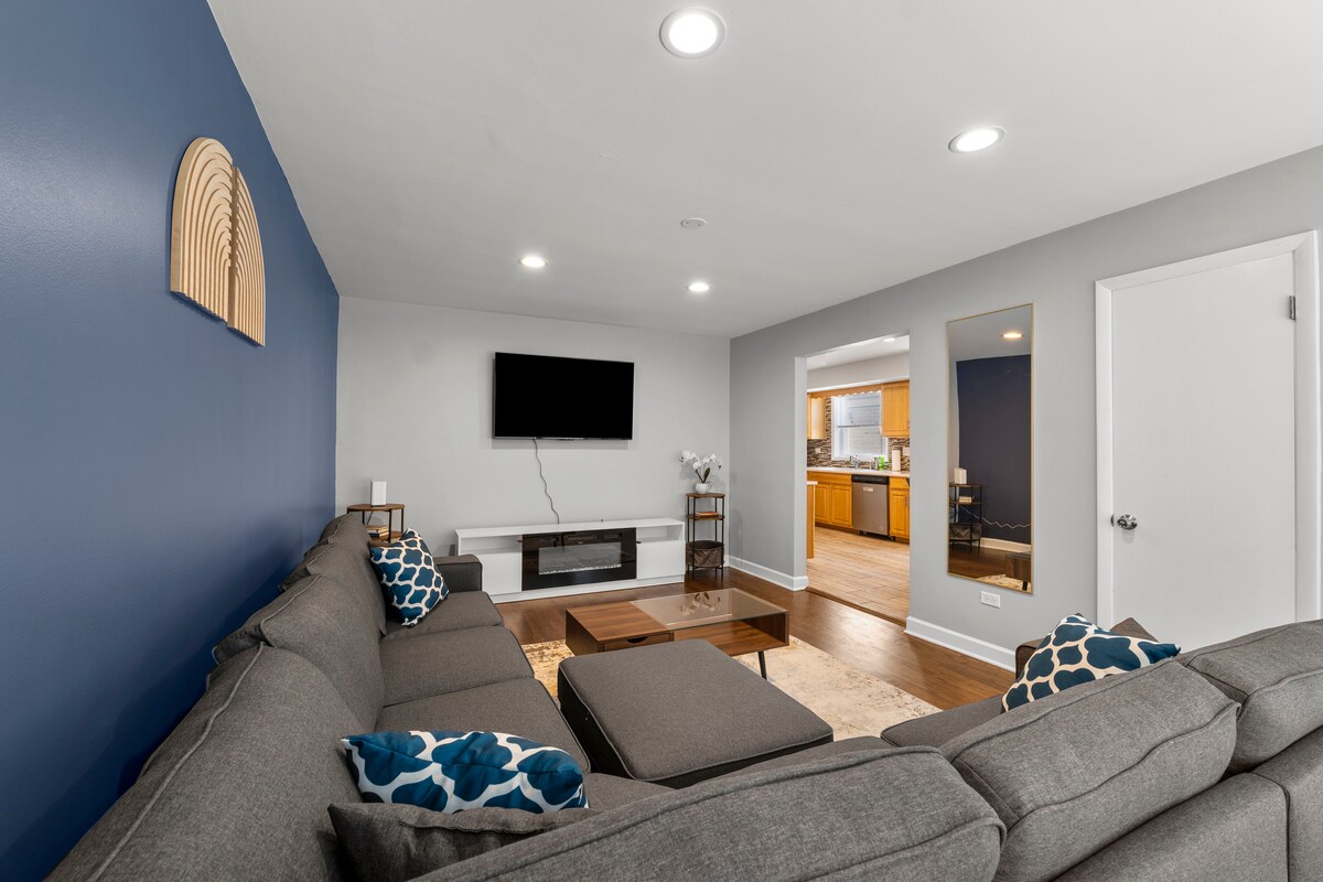 The living room is designed for comfort, featuring a gray sectional sofa adorned with decorative cushions. A coffee table sits in front, and a mounted 58-inch Smart TV is positioned on a wall, with a view into the adjacent kitchen area.