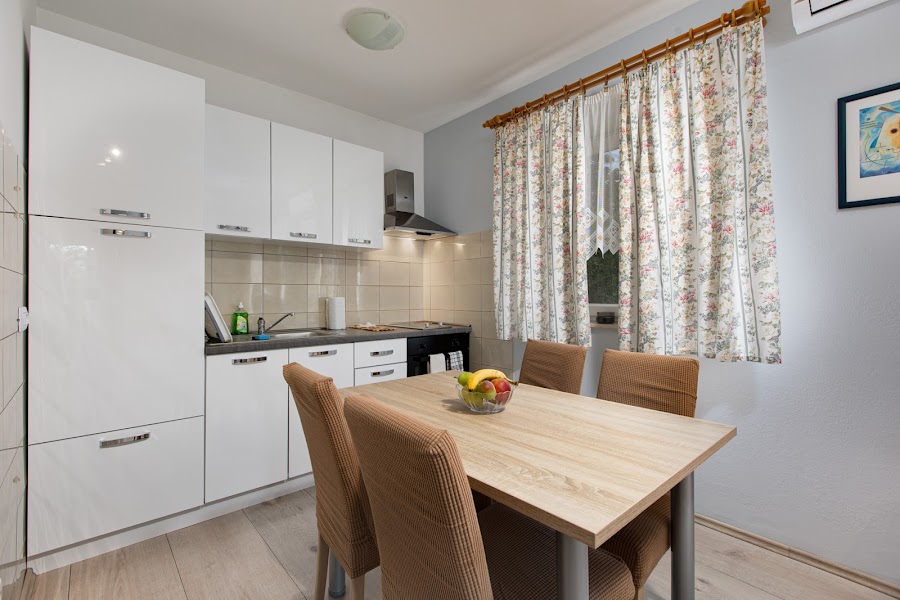 The kitchen is equipped with modern white cabinets and a countertop. A wooden dining table with four chairs is positioned at the center, featuring a bowl of fruit. Natural light filters through a floral curtain, enhancing the airy atmosphere.