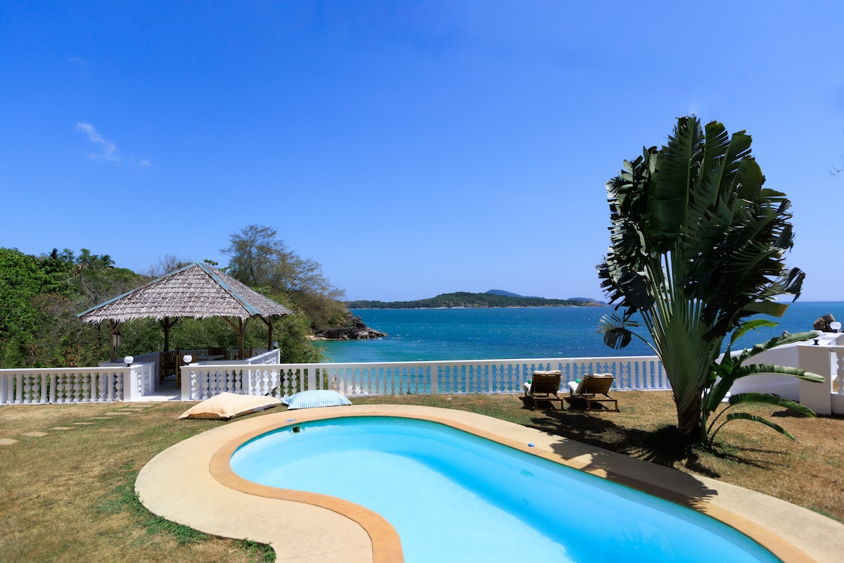 A private swimming pool reflects the bright blue sky, surrounded by a well-kept grassy area. Sun loungers positioned nearby invite relaxation, while a shaded gazebo provides a comfortable spot for gathering. A panoramic view of the sea can be seen beyond the pool.