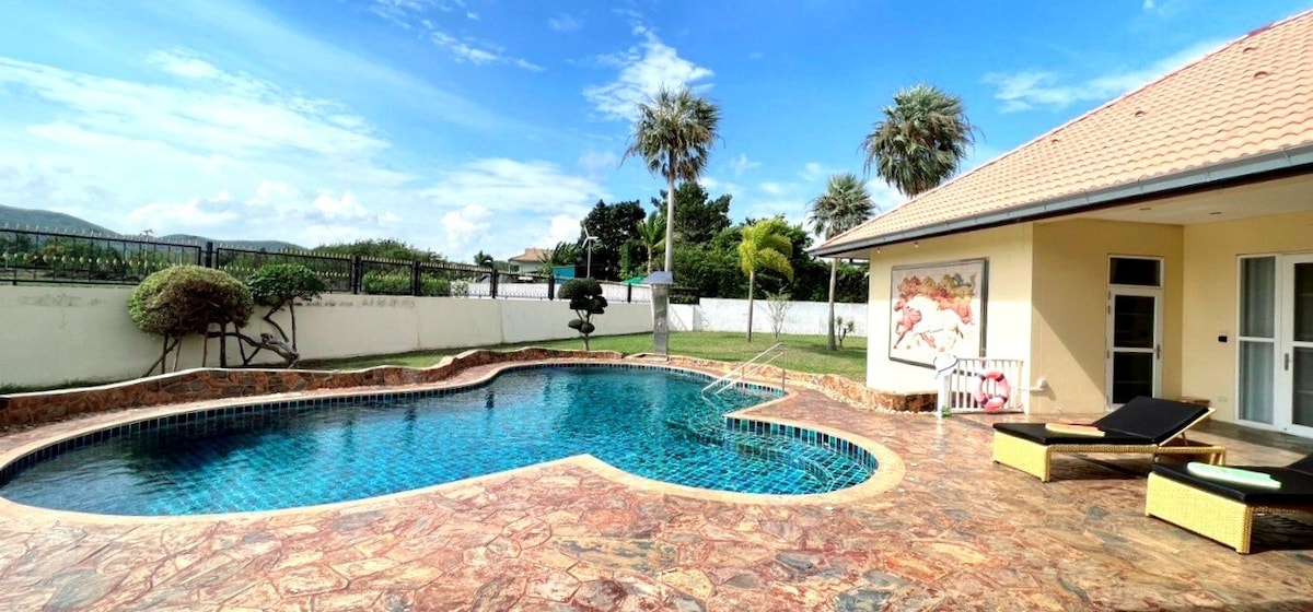 A private pool area features a curved design with clear blue water, surrounded by a sun-drenched terrace. Lounge chairs are positioned nearby for relaxation. Lush greenery provides a serene backdrop, while a mural adorns the exterior wall of the house, enhancing the inviting atmosphere.