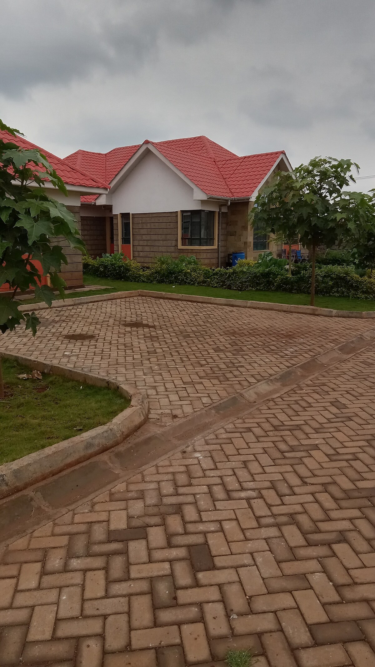A charming exterior of a single-story home is visible, featuring a red-tiled roof and stone façade. A well-maintained brick pathway leads to the entrance, surrounded by landscaped greenery and trees, offering a welcoming outdoor space.