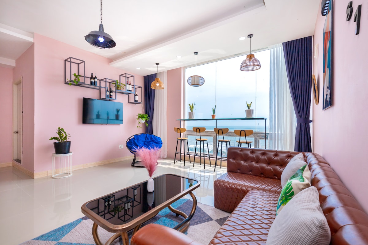 A cozy living area features a modern brown leather sofa and a sleek coffee table. A bar-style dining area with high stools and potted plants is visible against a large window, allowing natural light to illuminate the space. The walls are painted soft pink, creating a welcoming ambiance.