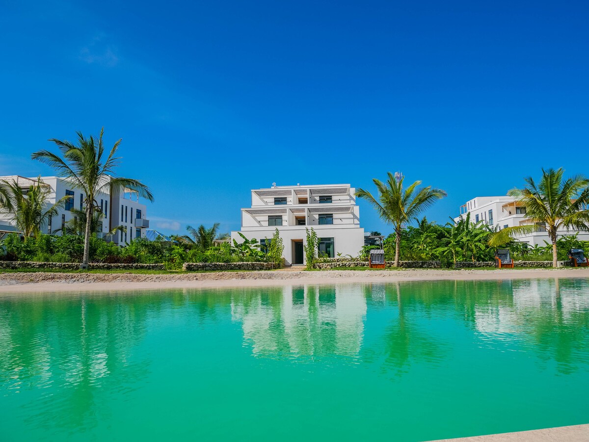 A modern building is reflected in a tranquil lagoon, surrounded by greenery and palm trees. The clear blue sky enhances the serene atmosphere of this residential leisure facility located along the East Coast of Zanzibar.
