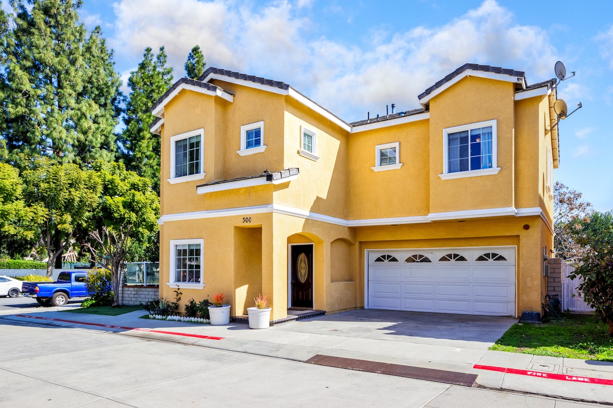 A two-story yellow house features a welcoming entryway with a circular window above a prominent front door. The driveway provides access to a two-car garage, surrounded by landscaped planters and a well-maintained exterior. Lush trees are visible in the background against a clear blue sky.