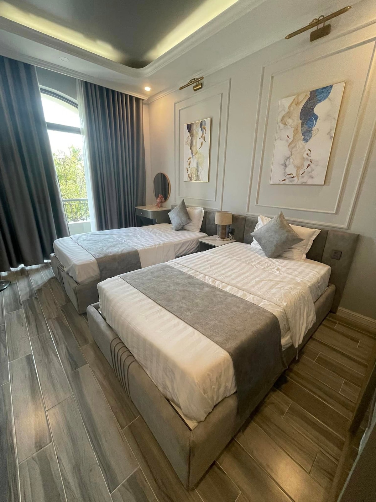A well-appointed sleeping area features two spacious beds, each adorned with light bedding and decorative pillows. Elegant artwork complements the neutral-toned walls. Natural light floods the room through large curtains, and a small bedside table with a lamp is positioned between the beds.