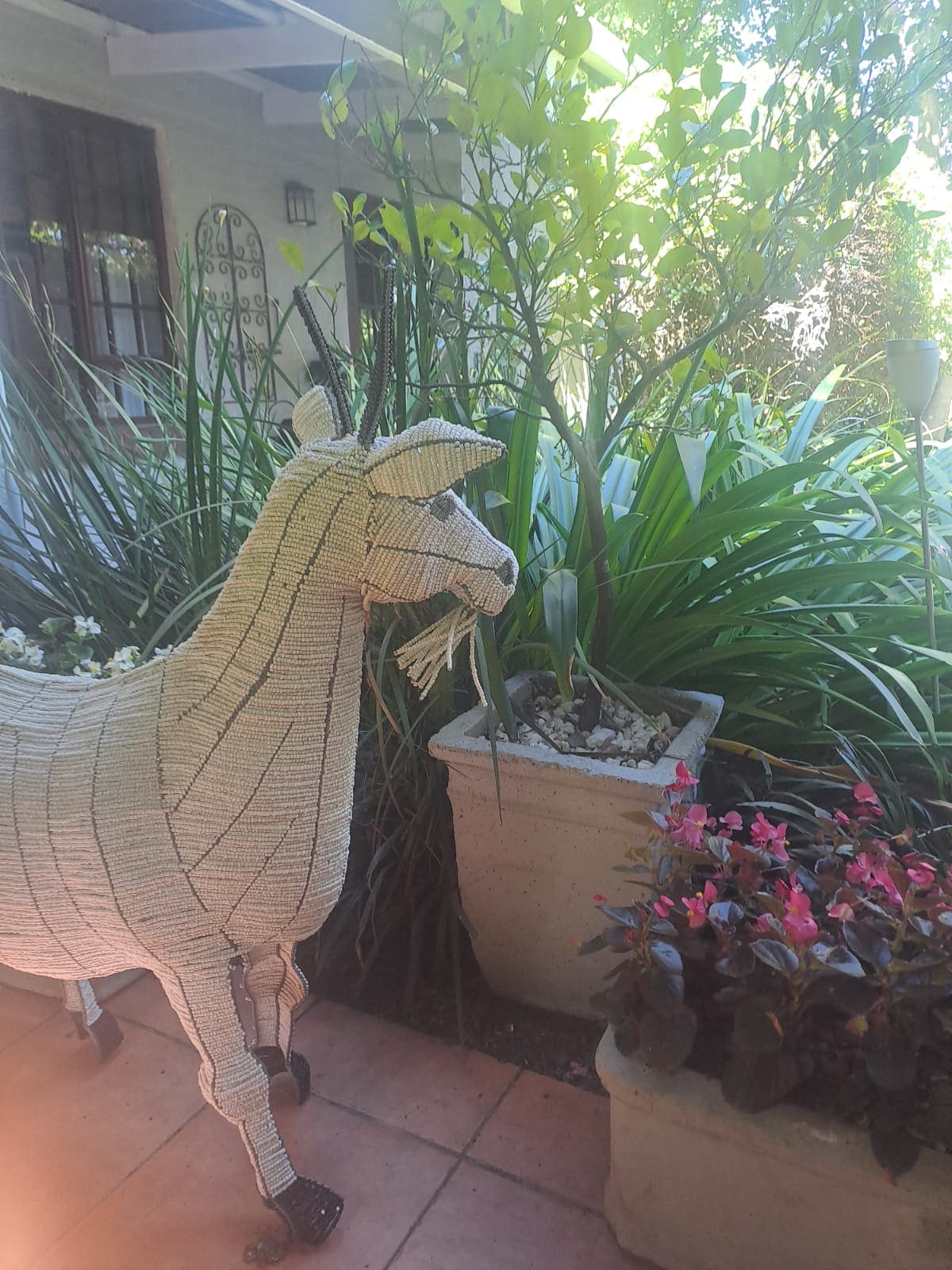 A charming woven goat sculpture stands among lush greenery and flowering plants. Surrounding the goat, potted plants feature vibrant foliage and blossoms, creating an inviting outdoor space. Natural light filters through the leaves, adding warmth to the serene garden setting.
