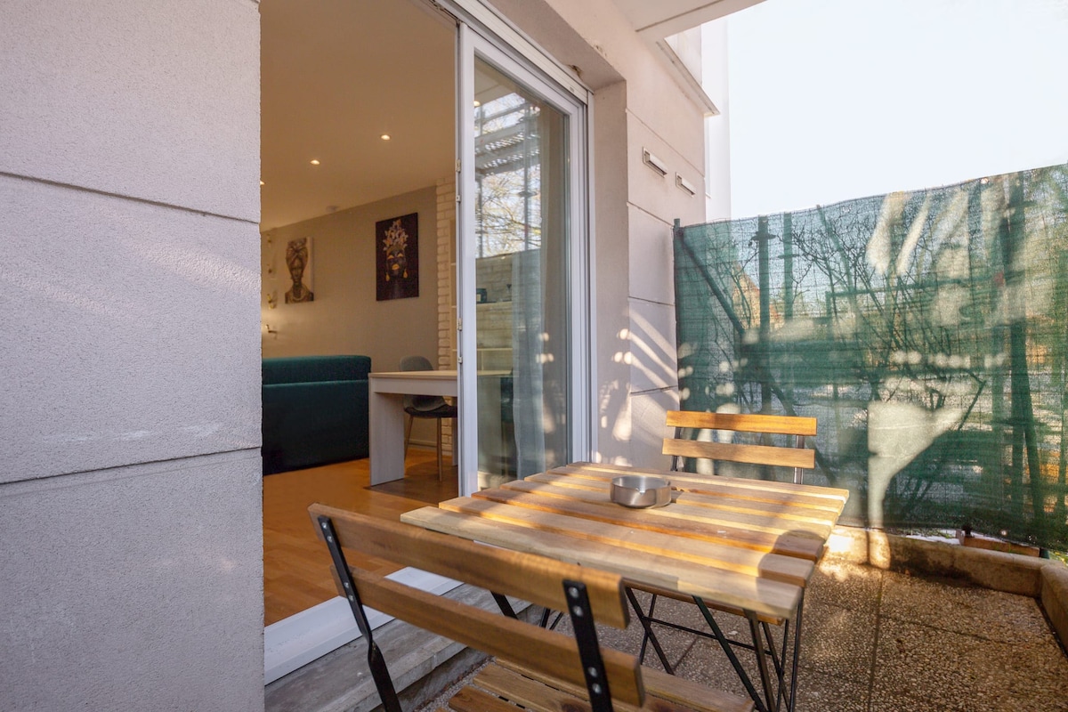An outdoor terrace is visible, featuring a wooden table with two folding chairs. Natural light illuminates the space, highlighting the texture of the table surface. In the background, an interior area is partially visible through glass doors, creating a seamless connection between indoor and outdoor areas.