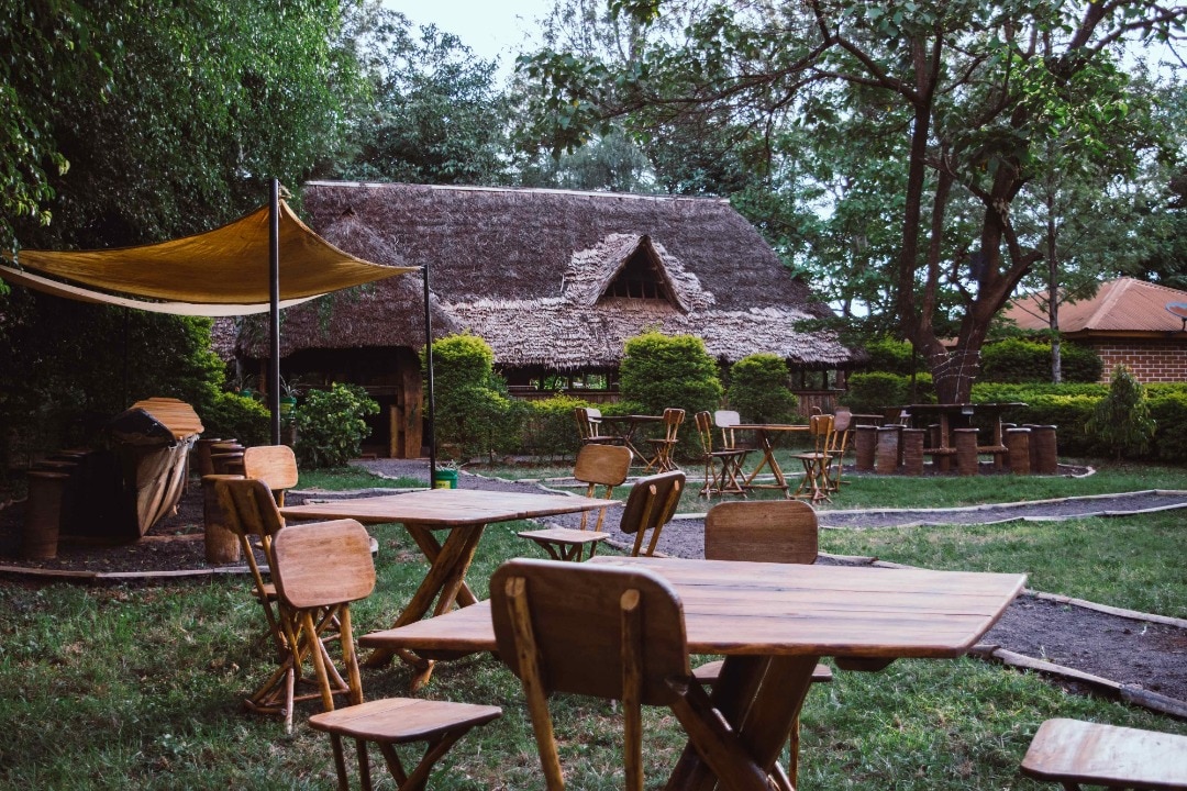 An outdoor seating area features wooden tables and chairs arranged on a grassy lawn. A thatched-roof structure is visible in the background, surrounded by manicured hedges and trees. A shade sail is positioned overhead, providing coverage from the sun.