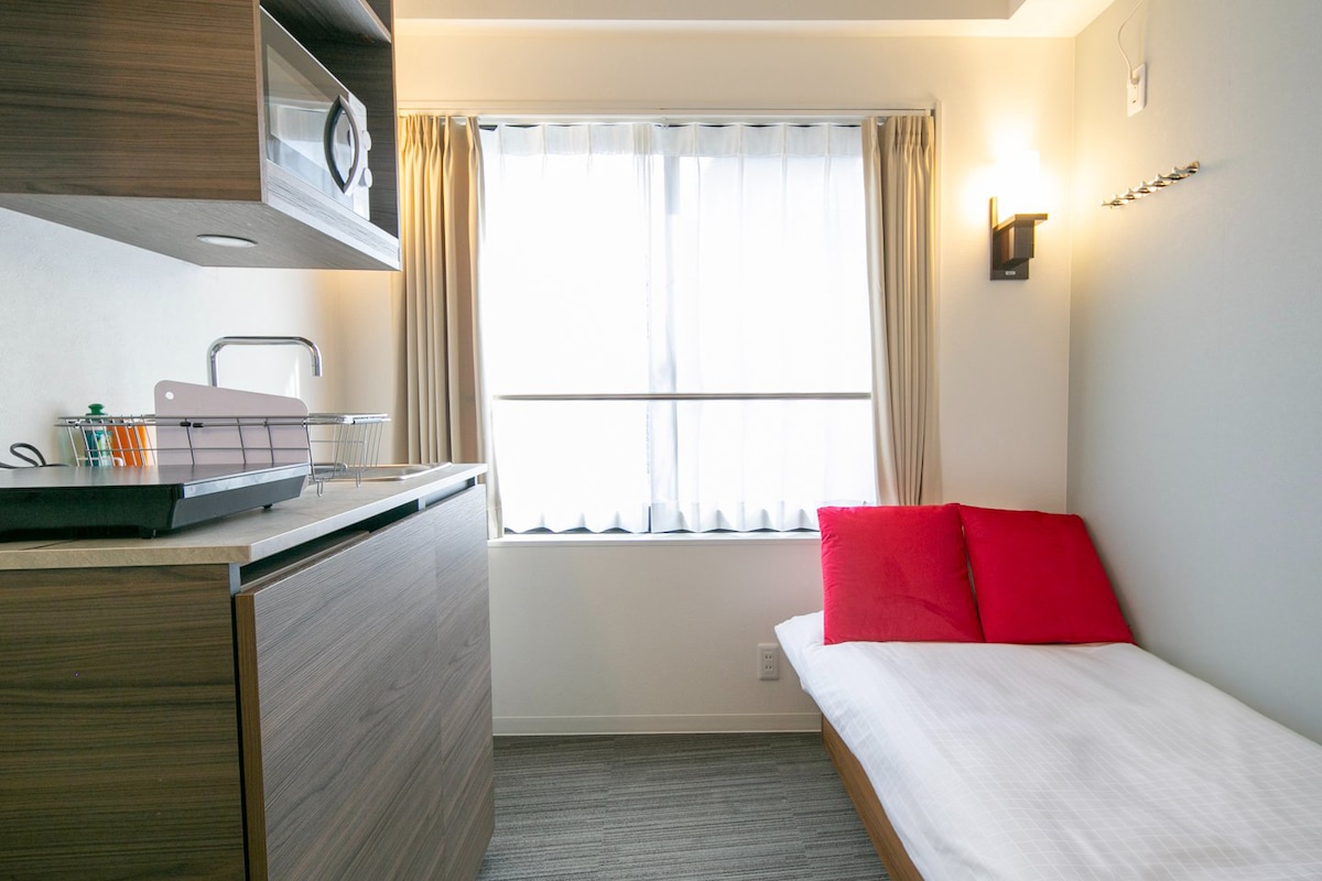 A tidy room features a single bed adorned with red pillows, positioned beside a window with sheer curtains allowing natural light. A compact kitchenette with wooden cabinets is seen along one wall, providing ample storage space.