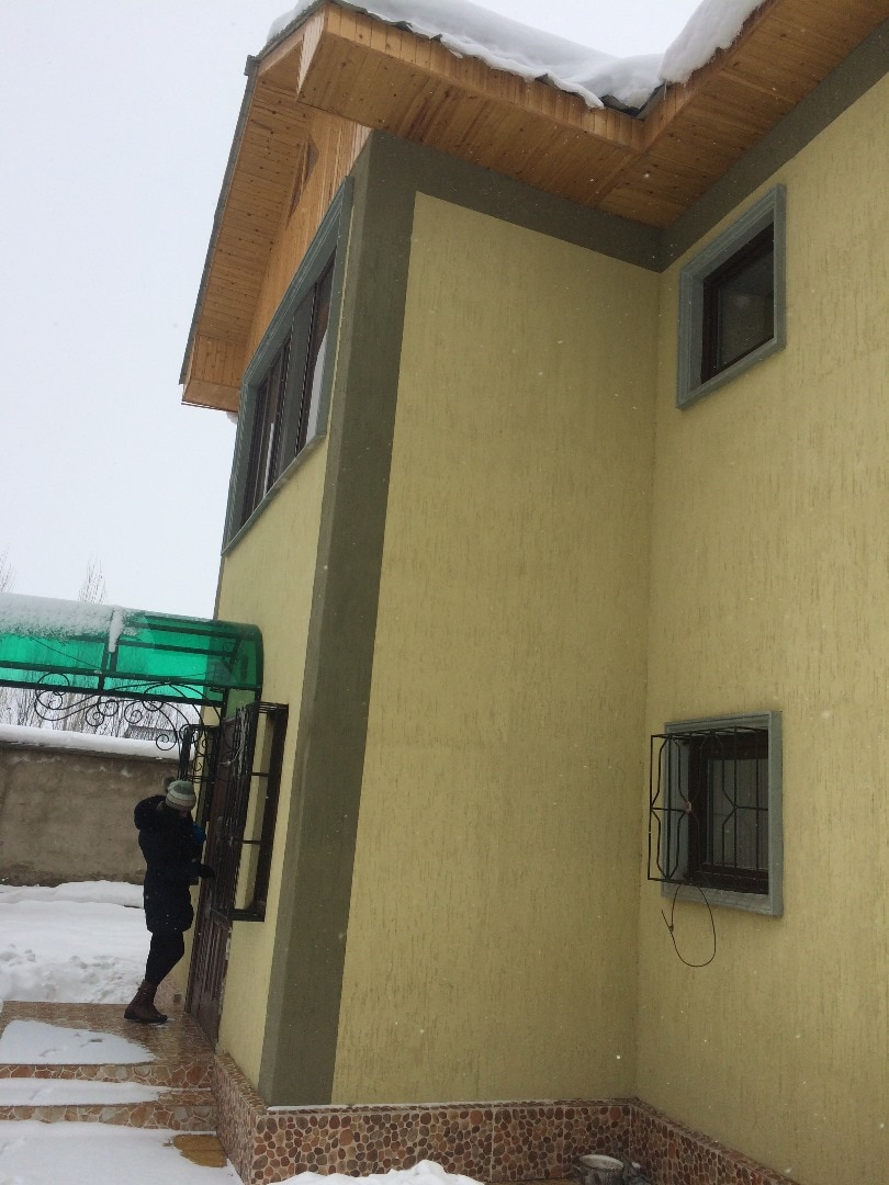 A corner of a two-story house is visible, featuring a light-colored exterior. Snow is gently falling, and a person is seen near a window, under a green canopy. Multiple windows can be observed, and the area around the house is covered in snow.