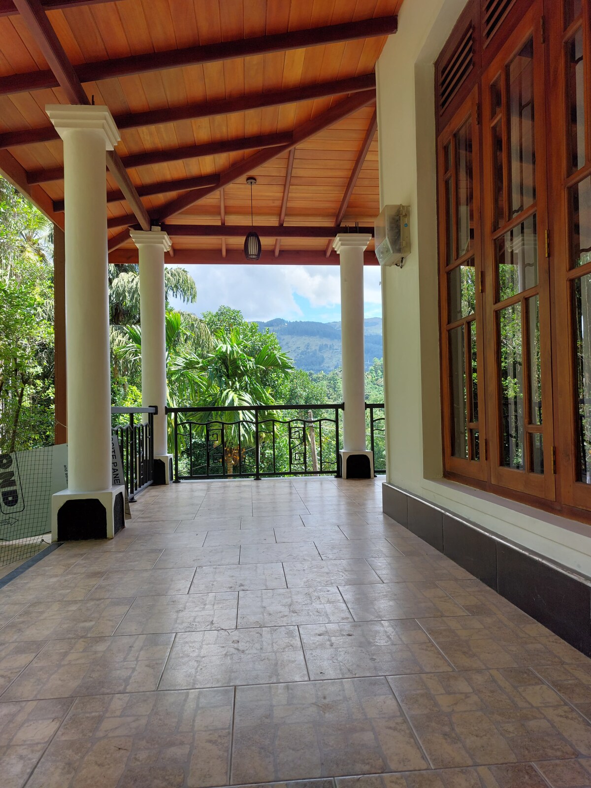 A spacious outdoor verandah is framed by tall wooden pillars and features tiled flooring. Large windows provide views of surrounding greenery and the distant mountains, enhancing the serene atmosphere. Natural light filters through, creating an inviting outdoor space perfect for relaxation.