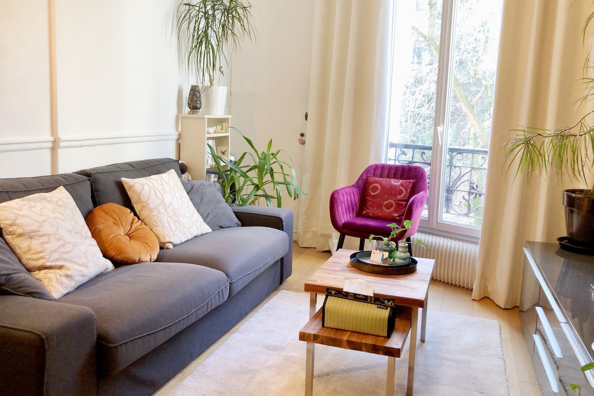 A comfortable living area features a grey sofa with decorative pillows, a vibrant purple armchair, and a wooden coffee table with potted plants. Large windows allow natural light to illuminate the space, framed by soft curtains, which enhance the room's serene atmosphere.