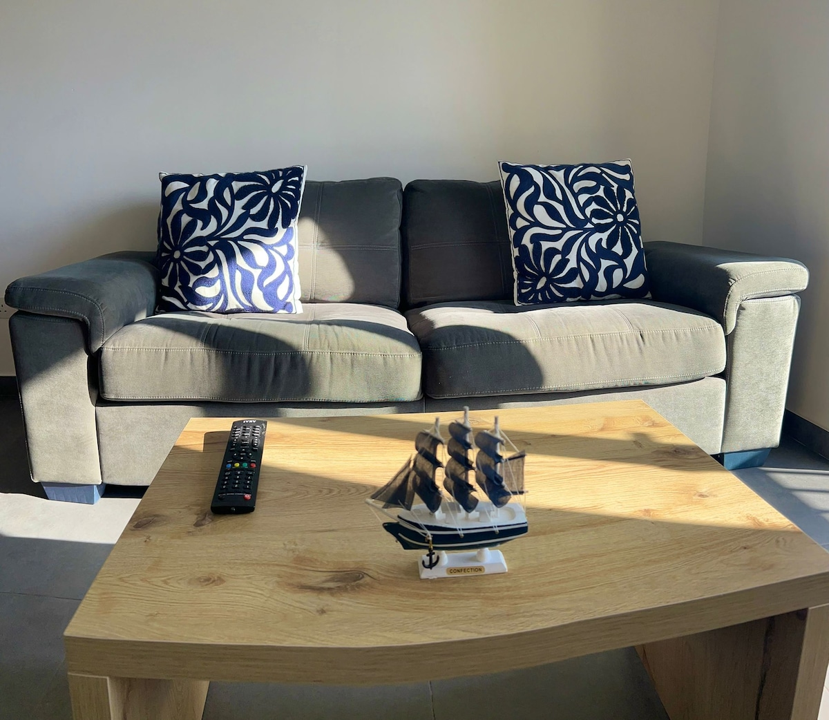 A comfortable sofa is situated in the living area, featuring two patterned cushions. A wooden coffee table is placed in front, holding a model ship. A remote control rests on the table's surface, with soft sunlight illuminating the space.