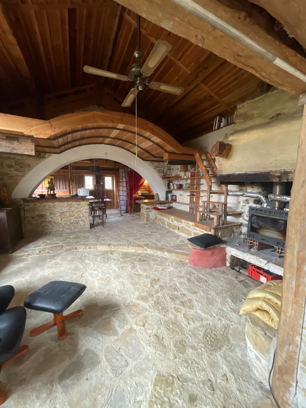 An open living area features natural stone flooring and a wooden ceiling with exposed beams. A curved archway leads to the kitchen space. A rustic fireplace is visible, along with various seating options, creating a cozy communal atmosphere.