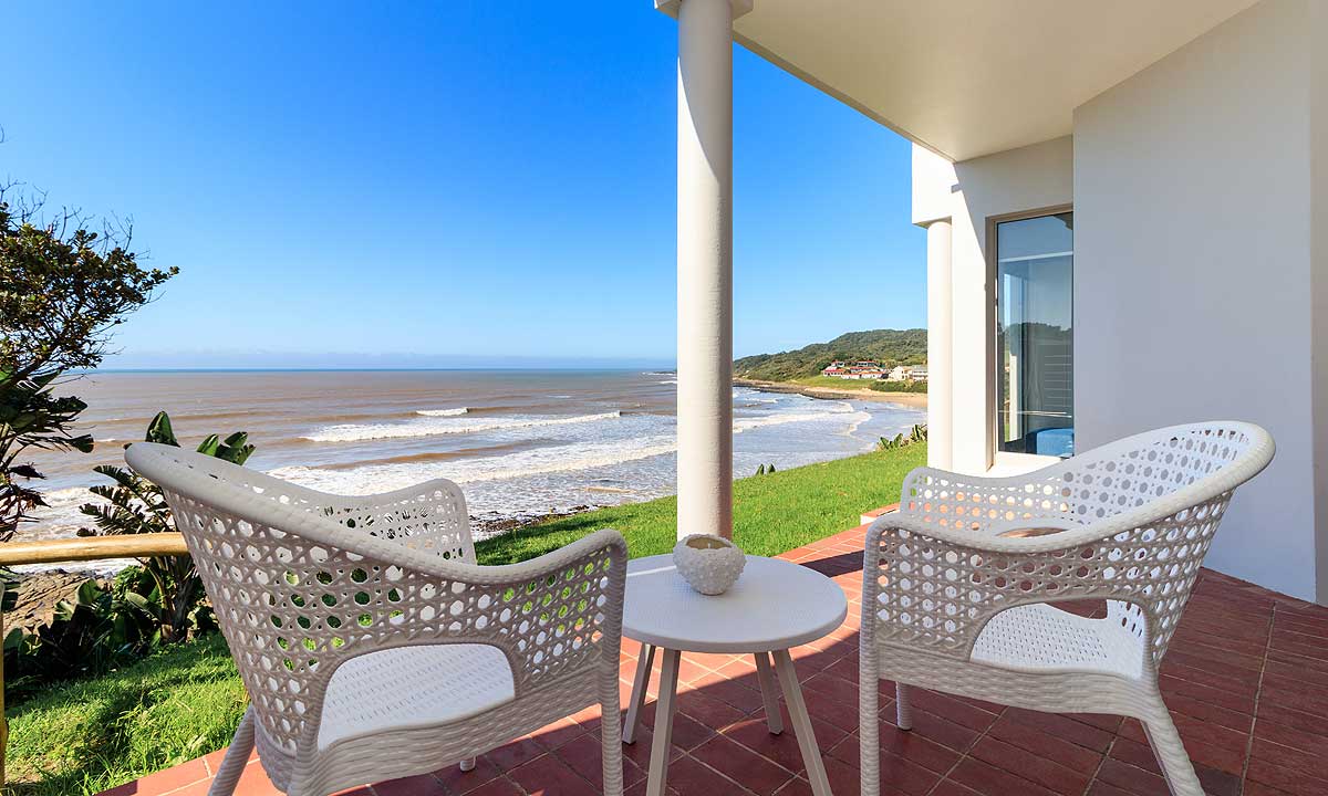 Two white wicker chairs are positioned around a small, circular table on a private veranda. The beachfront view showcases gentle waves lapping at the shore, complemented by a clear blue sky and gentle greenery in the foreground.