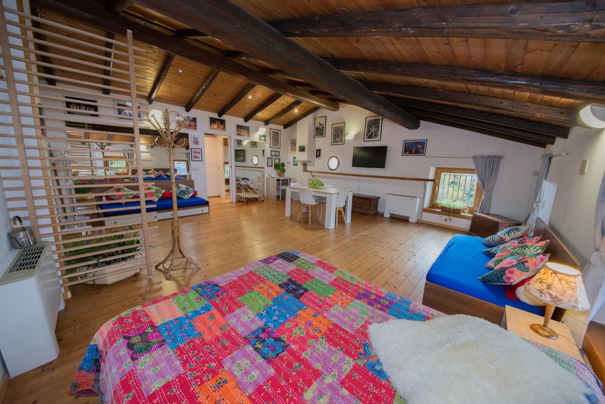An open-plan space is presented with wooden beams and polished floors. A vibrant quilt covers a spacious bed, while a dining area and workspace are visible in the background. Natural light filters through the windows, complemented by framed photographs on the walls.