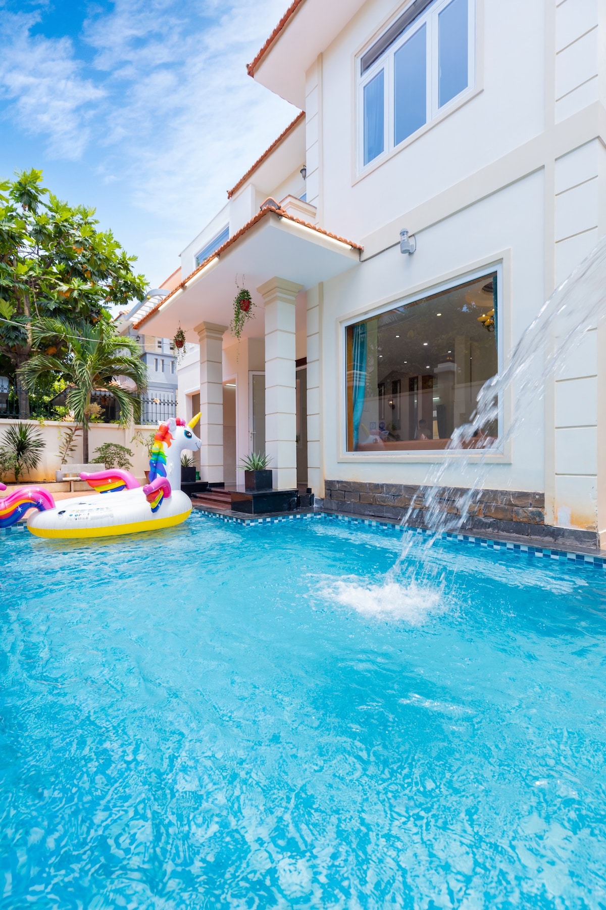 A spacious pool area features a refreshing blue pool with a waterfall element, surrounded by tropical greenery. The modern villa is partially visible, showcasing large windows that invite natural light. A colorful inflatable float adds a playful touch to the serene outdoor setting.