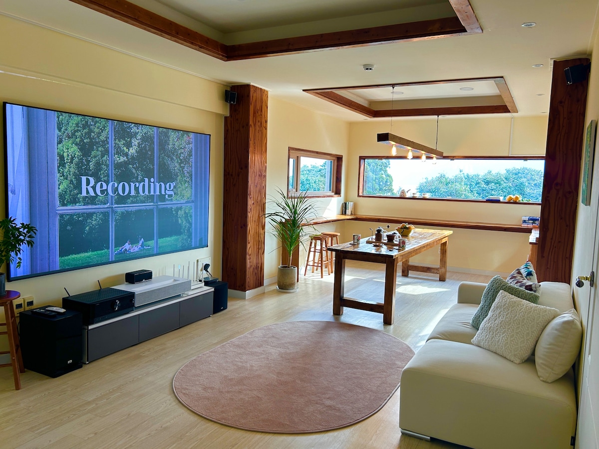 A spacious living area is seen, featuring a large screen displaying the word 'Recording.' A wooden dining table is visible, surrounded by chairs, alongside a cozy white sofa. Natural light filters through the large windows, illuminating the room.