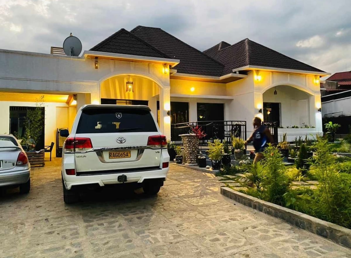 A spacious exterior view of Esspasso Home Boutique is presented, showcasing a well-maintained garden with various plants. A white SUV is parked in the driveway, while a pathway leads toward the entrance. Soft lighting highlights the architectural details of the house.