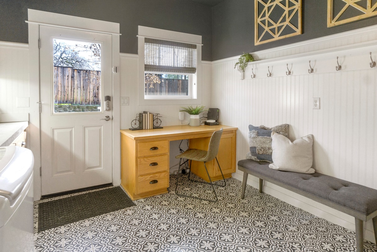 A functional laundry room is equipped with a front-loading washer and dryer. A wooden desk with drawers is positioned near a window, complemented by a chair. A bench offers seating next to decorative pillows, and a wall-mounted rack holds hooks for convenience.