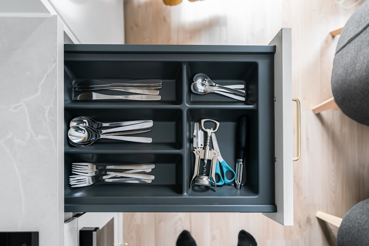 A kitchen drawer is shown, divided into compartments. Various utensils are neatly arranged, including silverware such as forks and spoons, a bottle opener, and a pair of scissors. The drawer's interior is dark gray with a light-colored exterior.