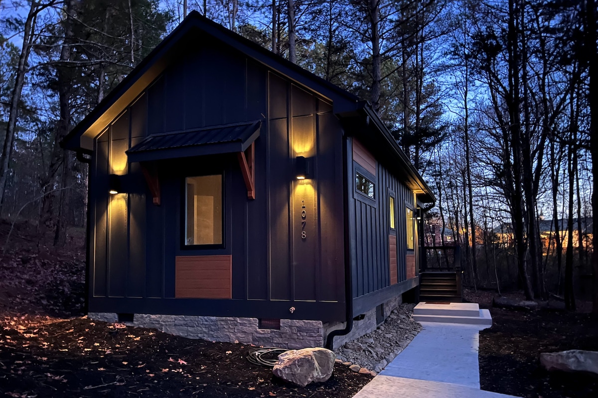 The modern cabin exterior is shown at dusk, featuring warm lights illuminating the dark wood siding. A welcoming pathway leads to the entrance, surrounded by natural landscaping with trees in the background, creating a serene atmosphere.