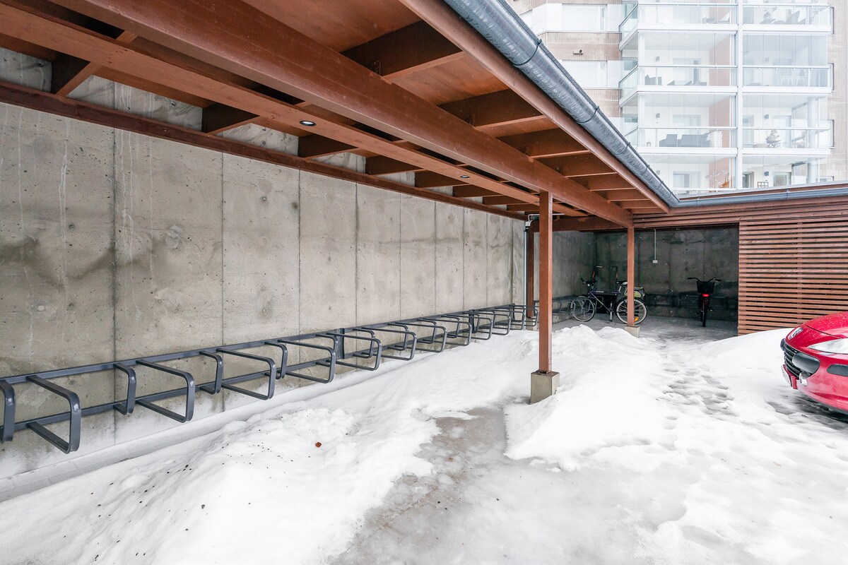 A designated parking area is visible, featuring a concrete wall and a wooden overhang. Bicycle racks are arranged along one side, with snow covering the ground. The structure includes a red car parked in a designated space, indicating accessibility for vehicle storage.